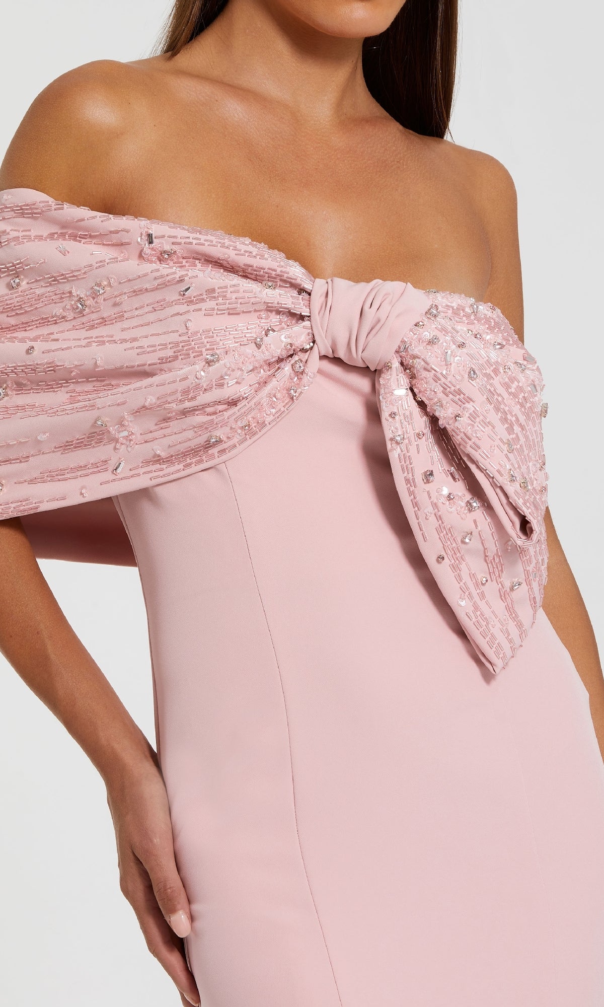 A woman wearing the Mac Duggal 2305 Draped Long Pink Formal Dress, featuring an off-the-shoulder design and a large sequined bow accent at the bust.